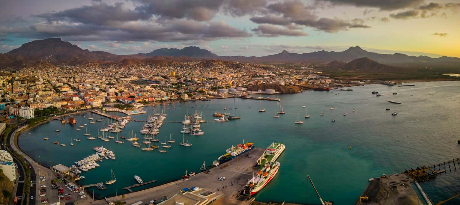 Panoramic aerial view of Mindelo city at sunset, with the marina and boats in the foreground, surrounded by the vibrant cityscape and mountains in the background under a warm, glowing sky