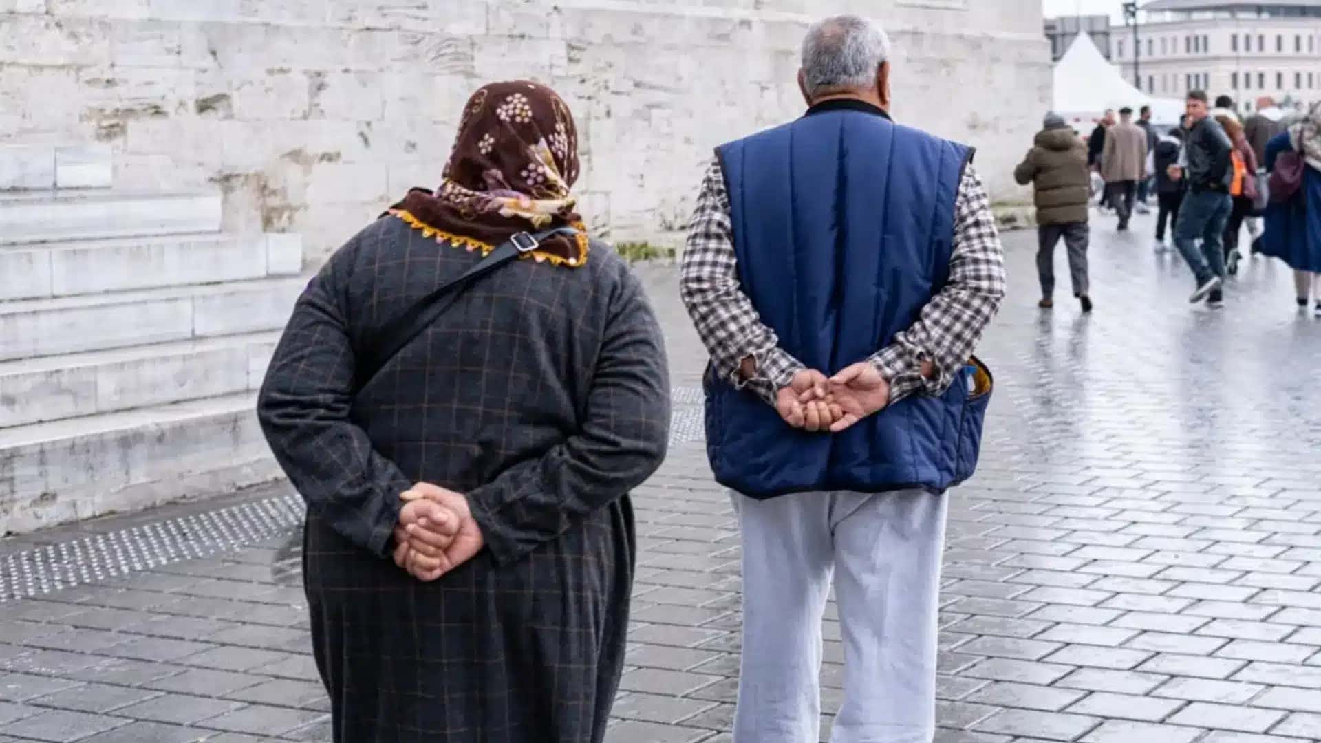 Duas pessoas caminham lado a lado com as mãos entrelaçadas atrás das costas em uma área pública.