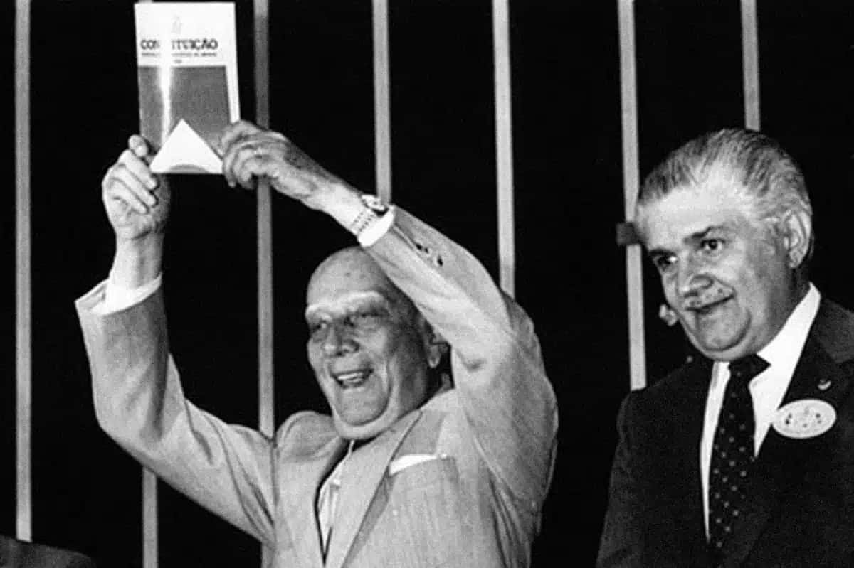Ulysses Guimarães, careca e sorridente, ergue a Constituição de 1988 com as duas mãos, ao lado de um homem de terno e gravata, também sorrindo, em um fundo escuro com listras verticais claras | Foto: Reprodução/Superinteressante