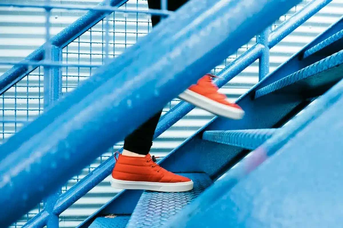 Pessoa subindo escada metálica azul, usando tênis acolchoado laranja com solado branco e calça preta. Gotas de água cobrem a estrutura da escada, sugerindo chuva ou umidade | Foto: Reprodução/Superinteressante
