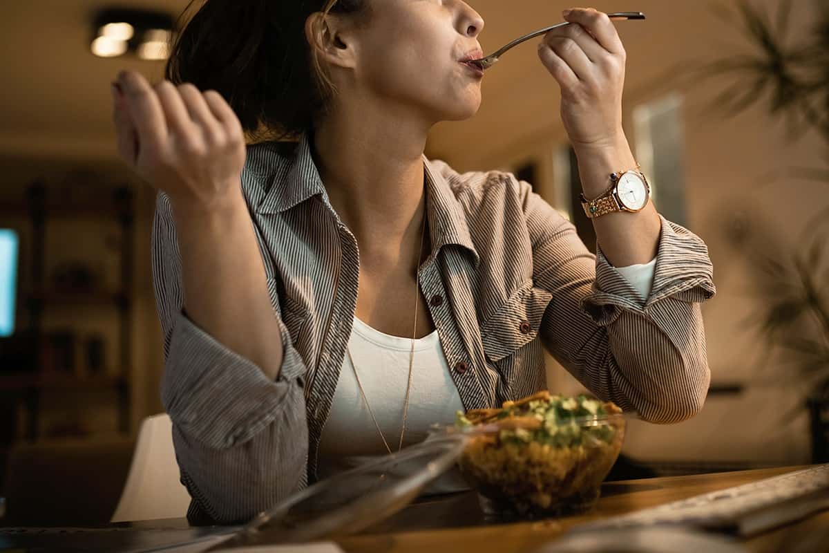 Low angle view of young woman eating salad with her eyes closed while using computer at night at home.
