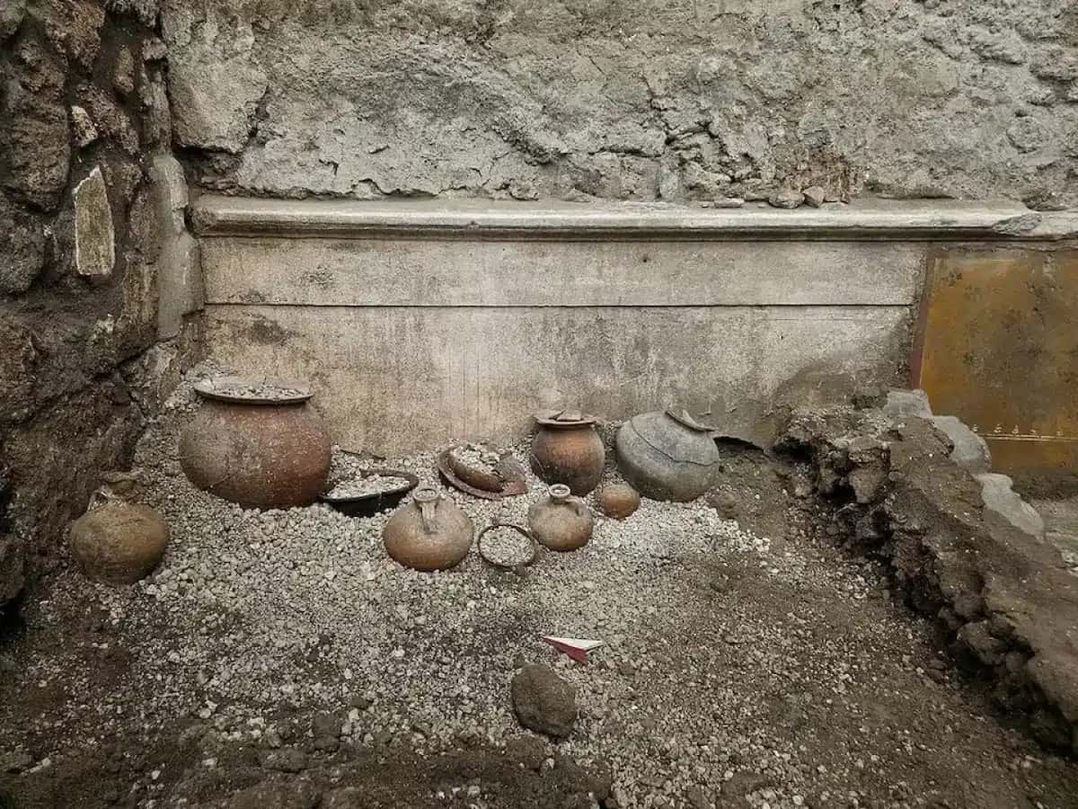 Várias ânforas e potes de cerâmica antigos, de diferentes tamanhos e tons terrosos, estão dispostos sobre um chão de cascalho claro, em um sítio arqueológico. Ao fundo, uma parede de pedra rústica e uma estrutura retangular de concreto aparente | Foto: Reprodução/Veja
