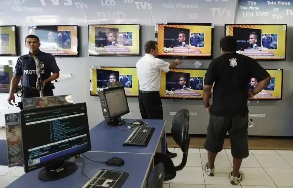 Membro da equipe de vendas mostra televisores de tela plana a um homem, enquanto ambos estão ao lado de um segurança do Bradesco, em uma loja da Casas Bahia em São Paulo.